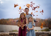 three children throwing leaves in the air in front of a brick wall